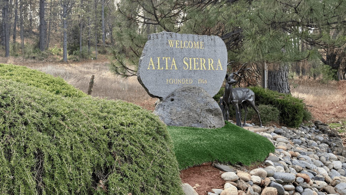 Welcome to Alta Sierra sign surrounded by pine trees, lush greenery, and a decorative deer statue, located in Nevada County, California